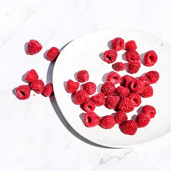 Vibrant red raspberries scattered on a white plate under bright daylight.
